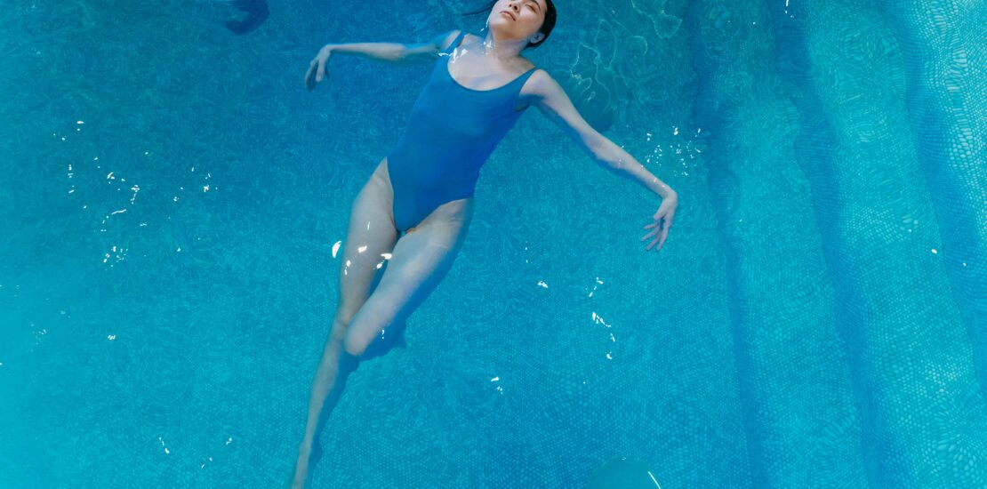 Image of an adult swimmer floating calmly in clear water — expression relaxed, bubbles rising, sunlight streaming through the surface.