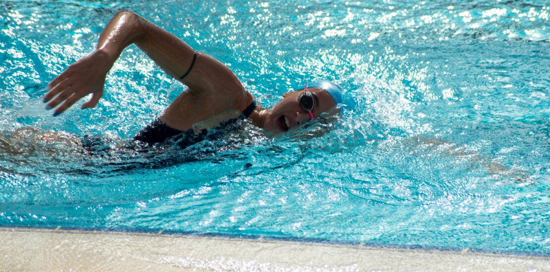 A swimmer wearing a blue swim cap and goggles performs freestyle in a clear blue pool. One arm is extended forward mid-stroke while the other pulls through the water. Sunlight reflects off the surface, highlighting the motion and energy of the swim. The image captures athletic technique and the dynamic flow of competitive swimming.