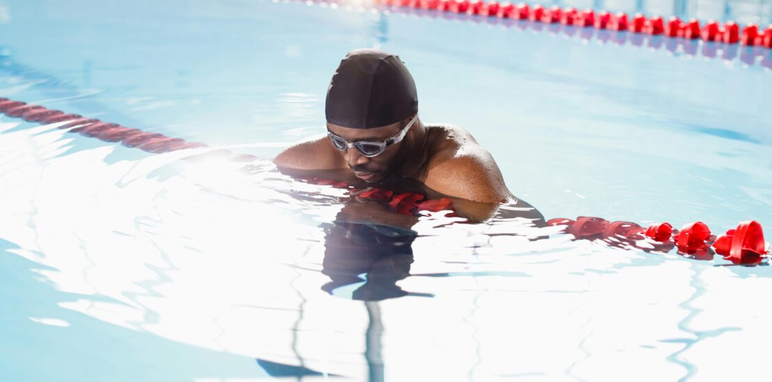 A swimmer wearing a black swim cap and goggles rests at the edge of a swimming lane divider in a pool. The lane markers feature red and green floating segments, and the clear water reflects light, creating a calm atmosphere. The swimmer appears to be catching their breath or pausing during training, highlighting a moment of focus and recovery in a competitive environment.