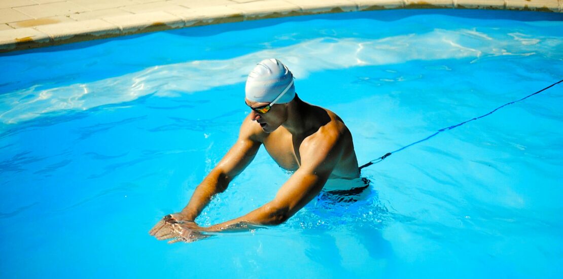 A swimmer in a bright blue pool wears a white swim cap and goggles while performing freestyle. They are tethered to the pool wall with a resistance cord attached at the waist, swimming powerfully against the tension. The taut cord highlights the strength and endurance training setup, with the vibrant water emphasizing motion and energy.