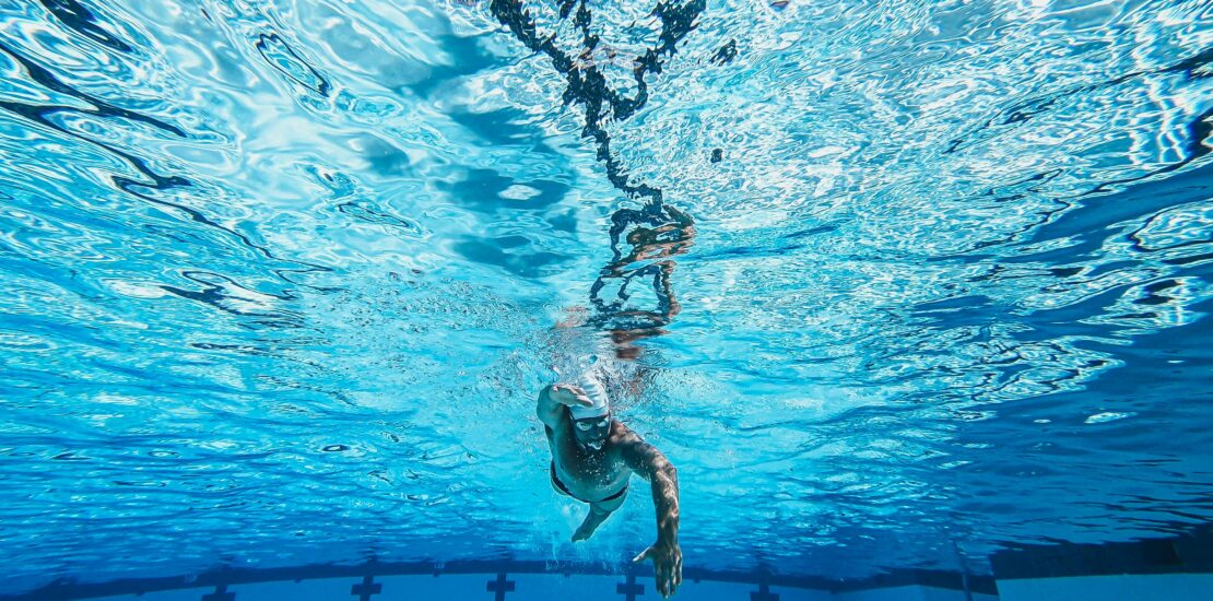 An underwater view of a swimmer performing freestyle in a pool. One arm is extended forward while the other pulls back through the water. Lane markings are visible on the pool floor, and crosses mark the side walls, indicating a competition or training pool. Sunlight filters through the surface, creating dynamic reflections and patterns across the clear blue water. The image highlights the swimmer’s form and fluid motion.