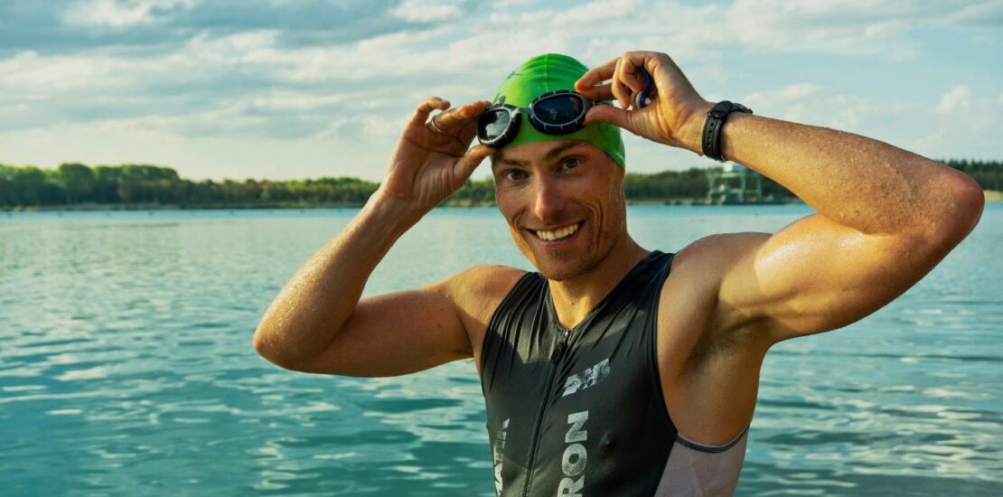 A swimmer stands smiling in front of a large lake with a forested shoreline in the background. They wear a green swim cap and hold goggles above their eyes, dressed in a sleeveless wetsuit with visible “LACRON” and “NEW BALANCE” branding. The bright daylight and partly cloudy sky highlight the outdoor, open-water setting, capturing a moment of preparation or celebration in a swim event.