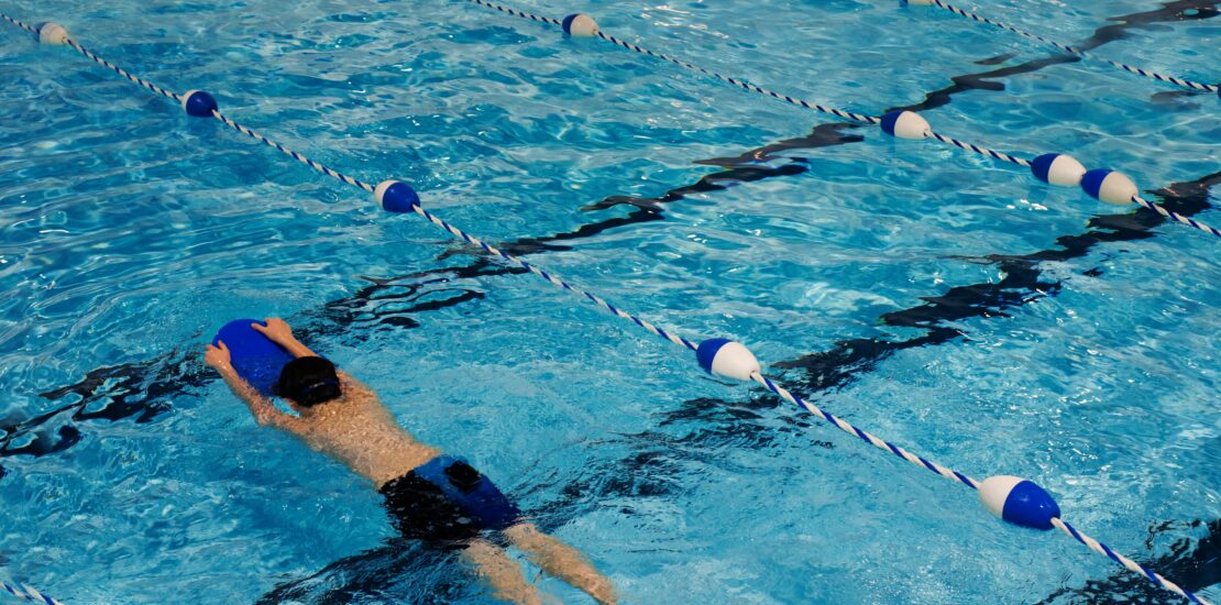 A swimmer practices in an indoor pool using a blue kickboard, gliding through a lane marked by blue and white dividers.