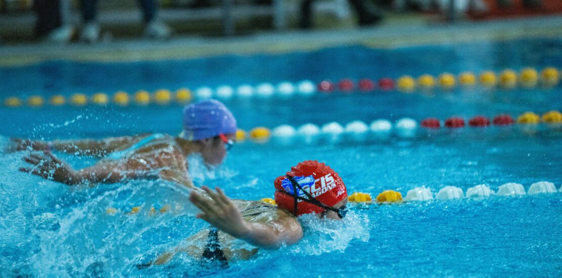 A swimmer wearing a red swim cap with the word and a shark logo performs a freestyle stroke in a clear blue pool. One arm is extended forward while the other is submerged, with the swimmer’s face just below the water surface. Lane dividers are visible on both sides of the pool.