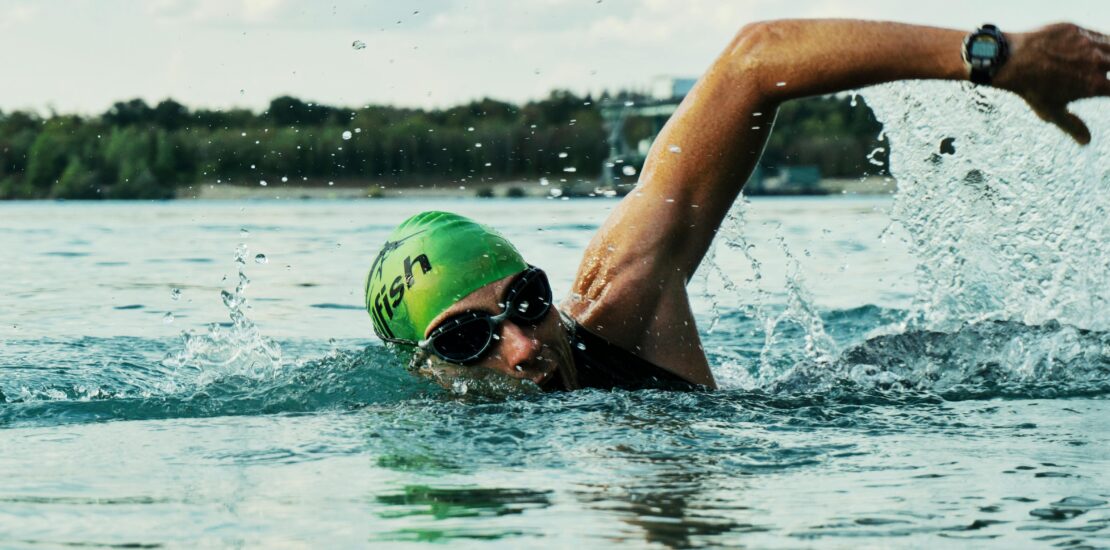 A swimmer performs freestyle in open water, wearing a green swim cap with the word “fish” printed on it and black goggles. Their right arm is mid-stroke above the surface, with droplets splashing around. A wristwatch is visible on their right wrist. In the background, a shoreline with trees and small structures is seen under daylight, highlighting the dynamic motion and intensity of outdoor swimming.