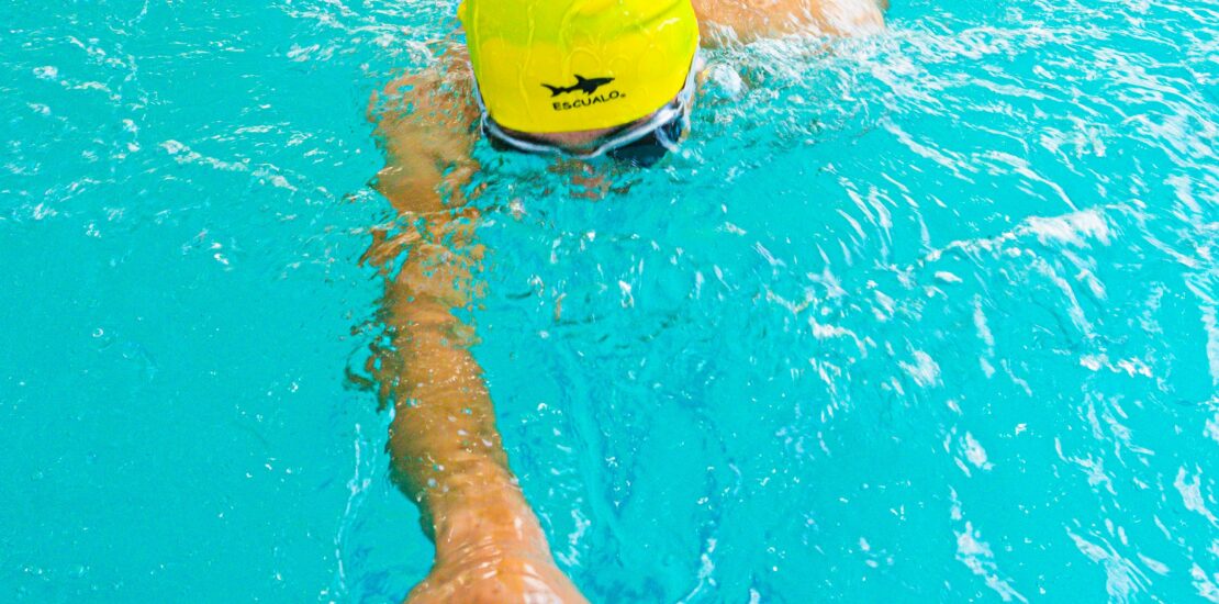 A swimmer wearing a bright yellow swim cap with the word “ESCUALO” and a shark logo performs a freestyle stroke in a clear blue pool. One arm is extended forward while the other is submerged, with the swimmer’s face just below the water surface. Lane dividers are visible on both sides of the pool.