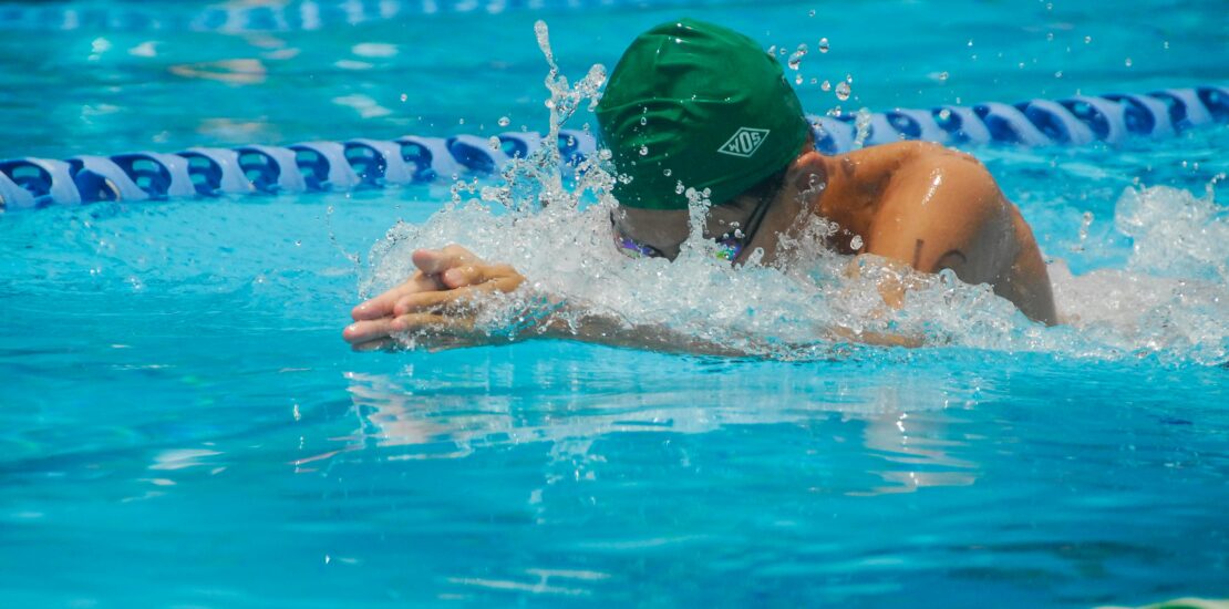A competitive swimmer wearing a green swim cap and goggles performs the breaststroke in a pool, arms extended forward with water splashing around, framed by blue and green lane dividers.