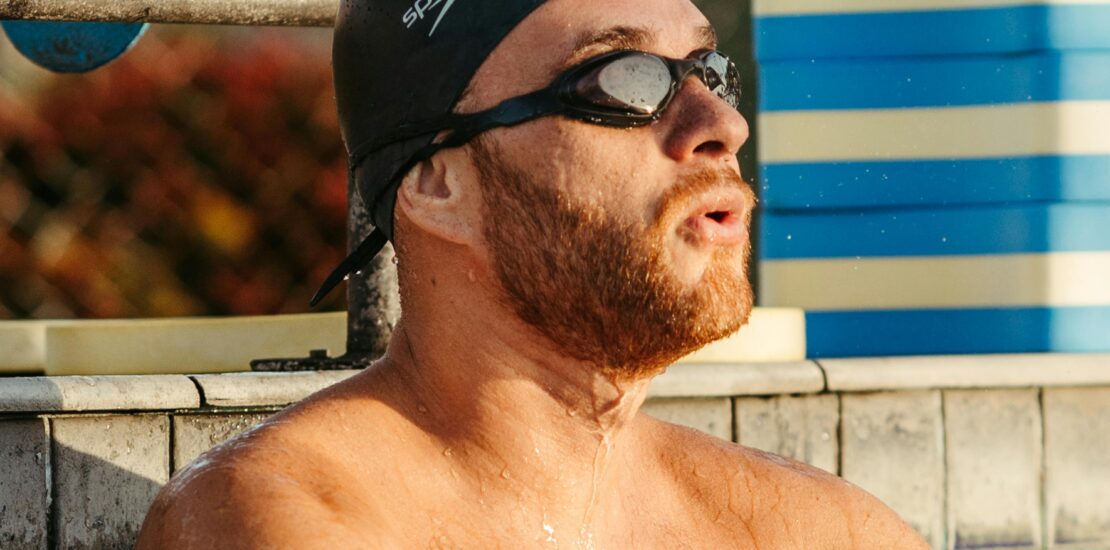 A swimmer wearing a black Speedo swim cap and dark goggles rests at the edge of a pool. Their shoulders and chest are visible above the water, with droplets on their skin. Behind them are a tiled pool wall, a metal railing, and stacked blue-and-white flotation devices, with a chain-link fence in the background.