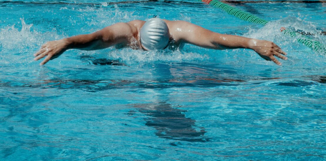 A swimmer wearing a light-colored cap and goggles performs the butterfly stroke in a swimming pool. Both arms are extended outward above the water, creating splashes. The pool has blue water with green and orange lane dividers, and diving platforms labeled “COLORADO” are visible in the background. Another person stands near the pool’s edge.