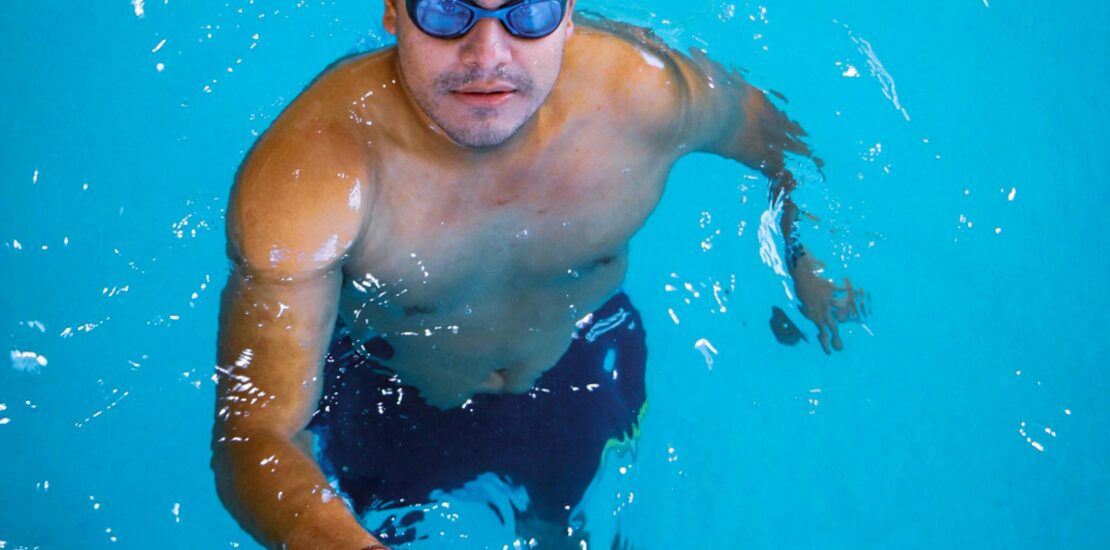 A man wearing a swim cap and goggles stands in a clear blue swimming pool, holding onto the pool edge and looking up at the camera.