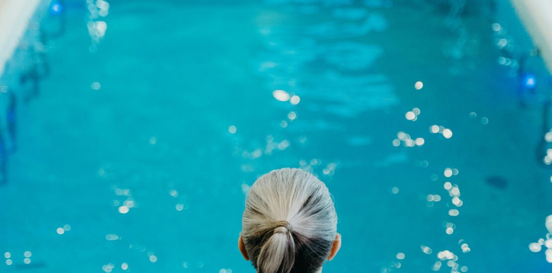 A person in a red swimsuit with gray hair tied back stands at the edge of a serene indoor swimming pool, preparing to enter the clear blue water. The background shows lounge chairs and tiled walls, creating a calm spa-like atmosphere.