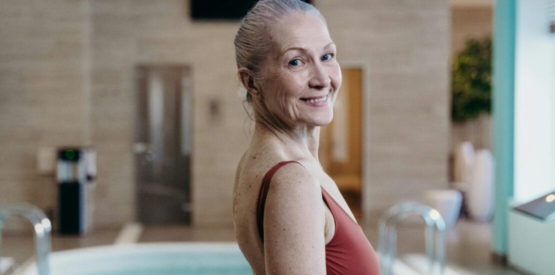 Older adult in a rust-colored swimsuit standing beside an indoor swimming pool with clear blue water, metal handrails, and beige tiled walls in a calm, well-lit leisure setting."