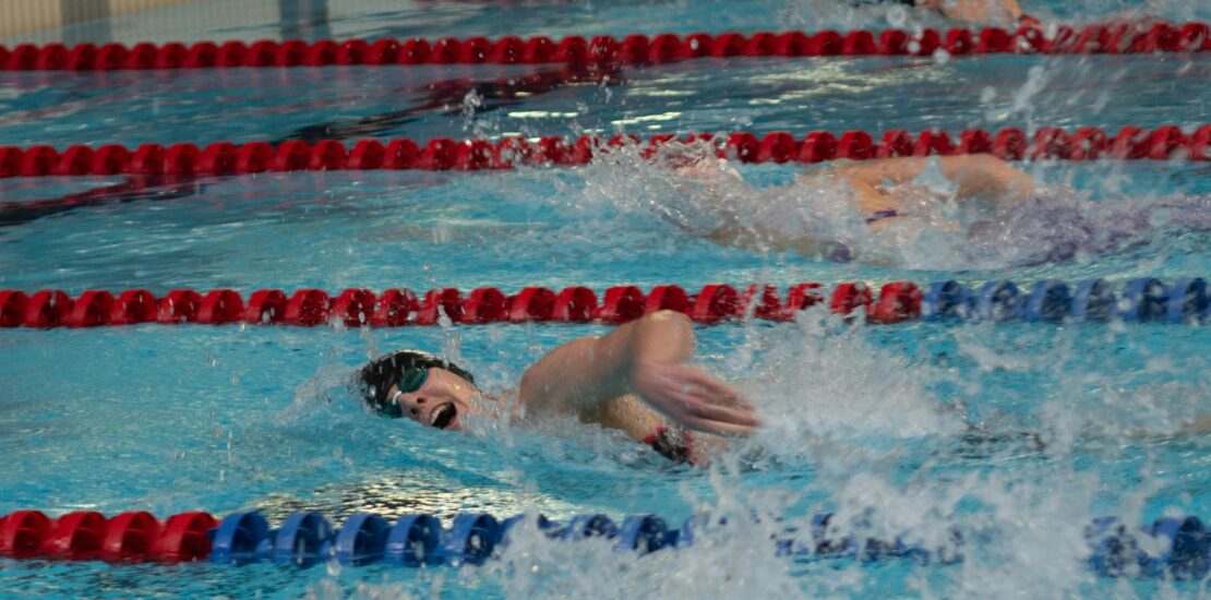 The image shows a swimming race in an indoor pool. Several swimmers are competing in separate lanes, divided by red and blue lane markers. The swimmer in the foreground is wearing goggles and a swim cap. He is performing the freestyle stroke, with one arm stretched forward and water splashing around him. The scene captures the energy, speed, and focus of the competition.