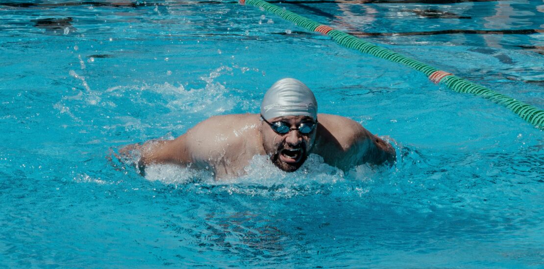 A male swimmer wearing a swim cap and goggles performs the butterfly stroke in a swimming pool, creating splashes as he moves forward in a marked lane.