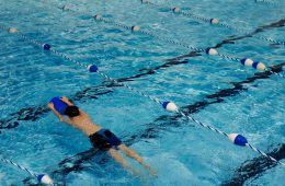A swimmer practices in an indoor pool using a blue kickboard, gliding through a lane marked by blue and white dividers.