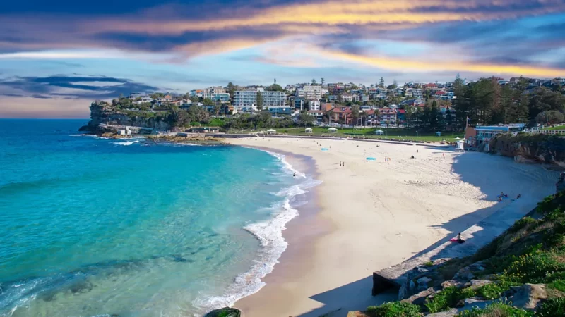 Coogee Beach in Sydney with turquoise waves, soft sand, and coastal homes under a vibrant sunset sky