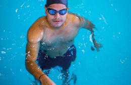 A man wearing a swim cap and goggles stands in a clear blue swimming pool, holding onto the pool edge and looking up at the camera.