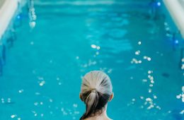 A person in a red swimsuit with gray hair tied back stands at the edge of a serene indoor swimming pool, preparing to enter the clear blue water. The background shows lounge chairs and tiled walls, creating a calm spa-like atmosphere.