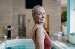 Older adult in a rust-colored swimsuit standing beside an indoor swimming pool with clear blue water, metal handrails, and beige tiled walls in a calm, well-lit leisure setting."