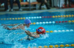A swimmer wearing a red swim cap with the word and a shark logo performs a freestyle stroke in a clear blue pool. One arm is extended forward while the other is submerged, with the swimmer’s face just below the water surface. Lane dividers are visible on both sides of the pool.