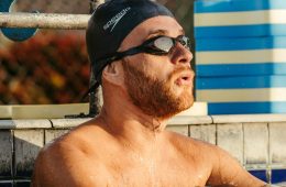 A swimmer wearing a black Speedo swim cap and dark goggles rests at the edge of a pool. Their shoulders and chest are visible above the water, with droplets on their skin. Behind them are a tiled pool wall, a metal railing, and stacked blue-and-white flotation devices, with a chain-link fence in the background.
