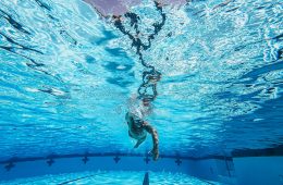 An underwater view of a swimmer performing freestyle in a pool. One arm is extended forward while the other pulls back through the water. Lane markings are visible on the pool floor, and crosses mark the side walls, indicating a competition or training pool. Sunlight filters through the surface, creating dynamic reflections and patterns across the clear blue water. The image highlights the swimmer’s form and fluid motion.