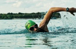 A swimmer performs freestyle in open water, wearing a green swim cap with the word “fish” printed on it and black goggles. Their right arm is mid-stroke above the surface, with droplets splashing around. A wristwatch is visible on their right wrist. In the background, a shoreline with trees and small structures is seen under daylight, highlighting the dynamic motion and intensity of outdoor swimming.