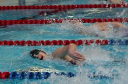 The image shows a swimming race in an indoor pool. Several swimmers are competing in separate lanes, divided by red and blue lane markers. The swimmer in the foreground is wearing goggles and a swim cap. He is performing the freestyle stroke, with one arm stretched forward and water splashing around him. The scene captures the energy, speed, and focus of the competition.