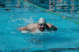 A male swimmer wearing a swim cap and goggles performs the butterfly stroke in a swimming pool, creating splashes as he moves forward in a marked lane.