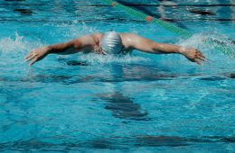 A swimmer wearing a light-colored cap and goggles performs the butterfly stroke in a swimming pool. Both arms are extended outward above the water, creating splashes. The pool has blue water with green and orange lane dividers, and diving platforms labeled “COLORADO” are visible in the background. Another person stands near the pool’s edge.