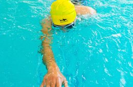 A swimmer wearing a bright yellow swim cap with the word “ESCUALO” and a shark logo performs a freestyle stroke in a clear blue pool. One arm is extended forward while the other is submerged, with the swimmer’s face just below the water surface. Lane dividers are visible on both sides of the pool.