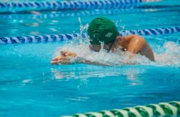 A competitive swimmer wearing a green swim cap and goggles performs the breaststroke in a pool, arms extended forward with water splashing around, framed by blue and green lane dividers.